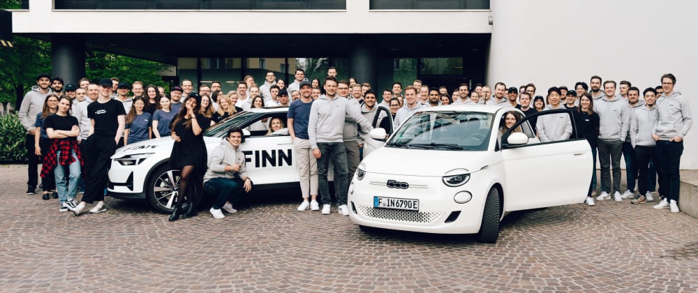 A group of around a hundred FINN employees poses with two cars in front of the FINN office in Munich, Germany
