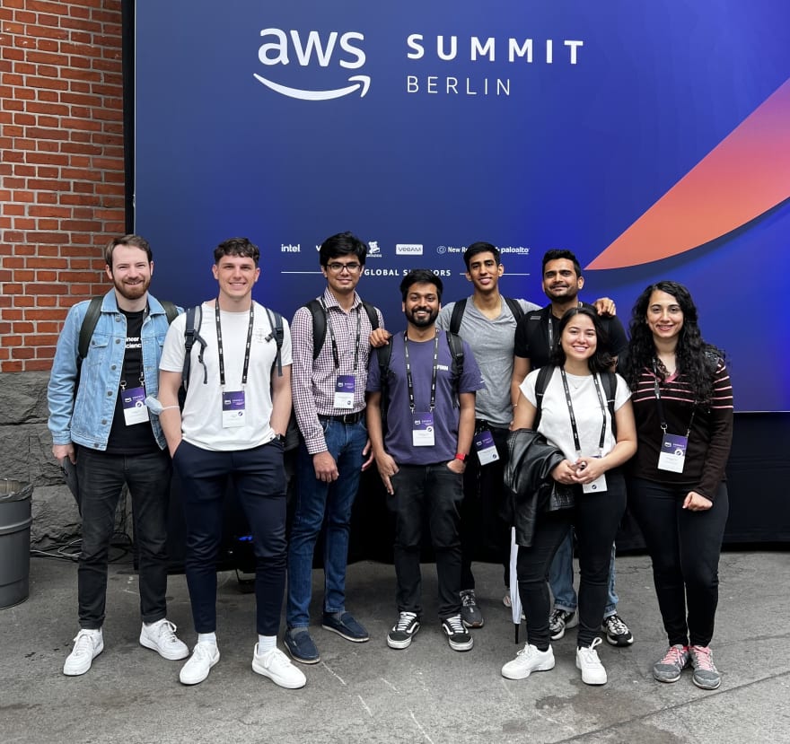 Eight people with conference badges standing in front of a huge sign with AWS Summit Berlin, smiling