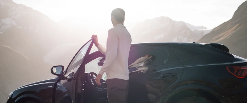 A person standing in sunlight next to a car, holding the car door, amid a vast mountainous landscape
