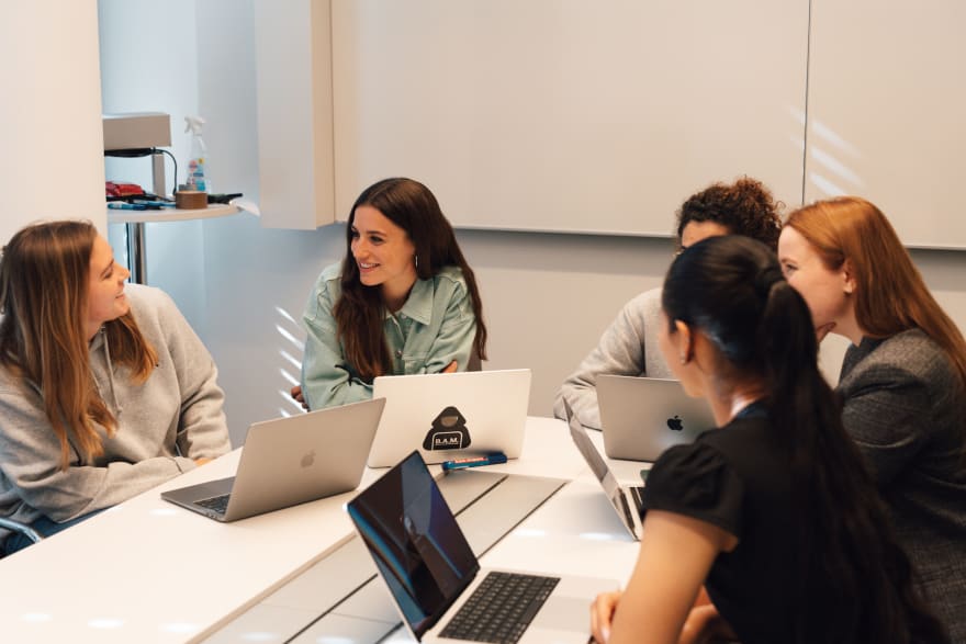 Five people sitting around a conference table with laptops, talking and smiling. One of the laptops has a sticker of a person in a hoodie with the words &lsquo;B.A.M.&rsquo; overlaid.